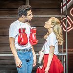 Danny (Marley Fenton) in jeans and white t-shirt, boxing gloves slung around his neck, stares down at Sandy (Hope Dawe) in Rydell gym top and skirt