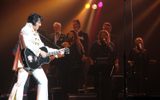 Shawn Klush in classic Elvis white high-collar suit, playing black acoustic guitar with singers behind him