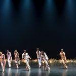 11 people in summer suits lean backwards from taking a step, long grass behind them