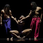 Two shirtless men in dreadlocks, left with purple trousers, right with red, hold their hands out to crouching woman who holds them with her arms crossed.