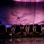 Dancers crouch holding a purple cloth billowing above their heads