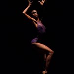 Black woman in purple leotard, hands thrown up, one leg bent up onto tip toes.