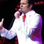 Ben Thompson as Elvis in white suit and red tie, singing into hand mic