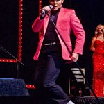 Ben Thompson as Elvis in red jacket, black top and trousers and white shoes, bending legs to the side as he sings into tilted standing mic