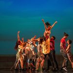 Group of dancers in summer-y outfits lift a woman in the air against dusky blue background