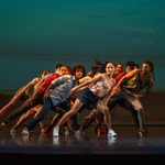 Group of dancers in summer-y clothes standing in pyramid formation lean to the left, hand balanced on leg in wide stance