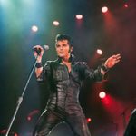 Dean Z as Elvis in all black leather, wide stance as he holds a standing mic