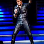 Dean Z as Elvis in all black leather, hand in the air as he sings into hand mic