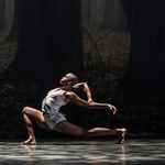 Black woman lunges deeply, grabbing the arm thrown straight out behind her on the dark stage