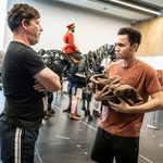 Haydn Burke unhappily clutches folden military jacket as Gareth Radcliffe, crossed arms, talks to him, puppet horses and cast in the background of the rehearsal studio