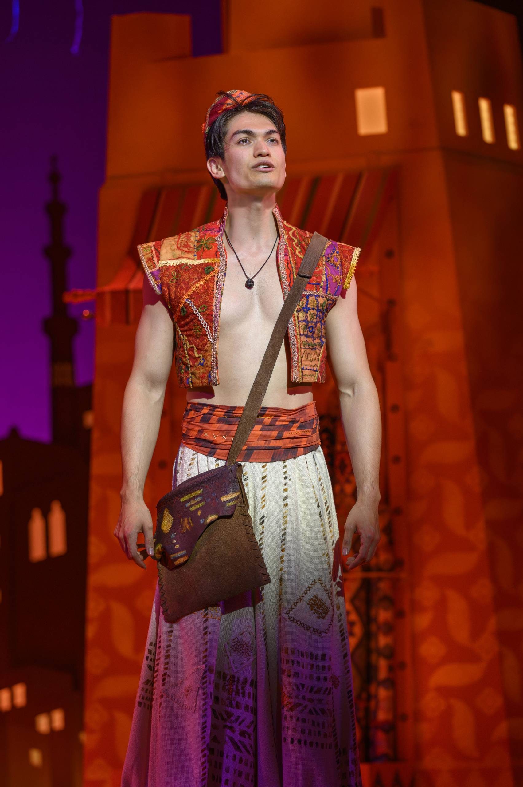 Gavin Adams as Aladdin in red waistcoat and fez and loose white trousers, leather satchel, looking up in Agrabah