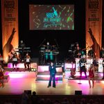 Fisher Stevens as Neil Diamond gestures to his left where the band and a blonde woman in red sparkly dress are, mirrored to his right are the orchestra and another woman