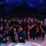 Fisher Stevens and the band and orchestra stand on stage, back to audience, posing for picture