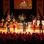 Fisher Stevens as Neil Diamond gestures to his right, where a blonde woman in red sparkly dress sings with a guitarist and the orchestra, mirrored to his left another woman, guitarist and the band