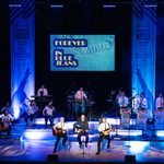 Forever in Blue Jeans written on screen above stage with band and orchestra wearing white, Fisher Stevens and two other guitarists out front