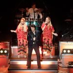 Fisher Stevens in black sparkly jacket, two blonde women in red dresses with palm trees on singing on the step behind him