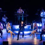 Fisher Stevens as Neil Diamond sitting on stall with two blonde women and two male guitarists