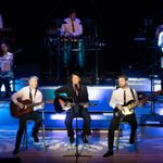 Fisher Stevens and two white men sit on stalls playing guitars