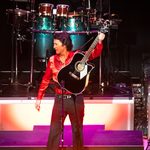 Fisher Stevens as Neil Diamond in red shirt holding up black acoustic guitar