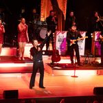 Fisher Stevens as Neil Diamond in black shirt, hand in the air, blonde women in red sparkly dresses singing behind, band playing to his left