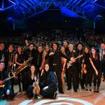 Fisher Stevens and the band and orchestra stand on stage, back to audience, posing for picture