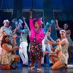 Oda Mae Brown (Jacqui Dubois) in bright pink jacket and large hat, raises a microphone up as ensemble dressed in money print jazzhands at her