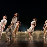 Seven dancers cross feet and lean back slightly, looking up. Long grass behind them.