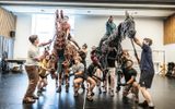 The cast of War Horse operating two life-sized puppet horses in rehearsal studio, riding towards the camera, brown horse on left with concerned rider on, black horse on right with rider holding out pretend weapon