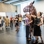 Tom Sturgess patting front flank of brown puppet horse as Alexandra Donnachie reaches up to the head whilst operating it via long stick, cast watching on.