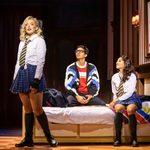 Maia Hawkins in school uniform standing at the end of a bed which Luke Latchman and Nikita Johal are sitting on