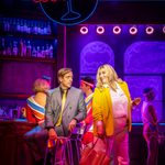 Chris Grahamson in grey suit and gold tie turns around in his bar stool to look at Melissa Jacques in yellow suit, under Cocktail neon sign