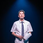 Evan (Ryan Kopel) in school shirt and tie and arm cast reading Connor, looking up under single spotlight