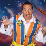 Silly Simon (Kev Orkian) pulling a wide face and jazz hands, wearing blue, yellow and orange striped waistcoat and white shirt