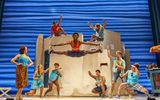 Black man does jumping splits as Tanya watches in bathing suit and youngsters cheer him on, some from the rooftops of greek building