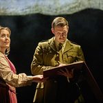 The Singer (Sally Swanson) holds a book as Lieutenant Nicholls (Chris Williams) reads it.