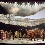 Joey (puppeteers Matthew Lawrence, Rafe Young, Felicity Donnelly) pulls a plow as crowds watch below a banner like ripped paper with pencil drawing of countryside on