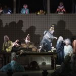 Rigoletto (Daniel Luis de Vicente) points down from a table at a debaucherous party at a man with a long beard and black robe