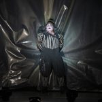 Rigoletto (Daniel Luis de Vicente) in black and white striped top and leggings with black trousers and pointed hat, clutches the grey hanging behind him as he backs away, mouth and eyes wide in fear