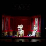 Maddalena (Alyona Abramova) sit backwards in a chair, The Duke (Raffaele Abete collapsed against the red and mirrored wall as Gilda (Soraya Mafi) and Rigoletto (Daniel Luis de Vicente) look in from the shadows outside