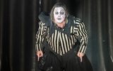 Rigoletto (Daniel Luis de Vicente) in black and white stripe top and black ruffle, with white face paint and exaggerated eyebrows leans forward, frowning and pointing downwards with both hands