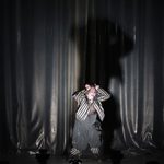 Rigoletto (Daniel Luis de Vicente) pointing his fingers on his head like horns, his shadow looming behind him, his white and black striped jacket open