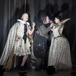 Two white wearing courtiers (Nathanaël Tavernier and Alyona Abramova) menace Rigoletto (Daniel Luis de Vicente) who peers deeply into the one to his left's face