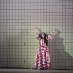 Gilda (Soraya Mafi) in red print dress, leans against wall, arms thrown wide above her