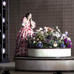 Gilda (Soraya Mafi) in red print dress clutches her face in pleasure as she stands next to round flowerbed