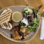 A panini on a brown oval plate with lettuce, vegetable crisps and a small pot of salad dressing. Next to the plate is a knife and fork wrapped in a napkin.