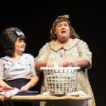 Edna sorts laundry in a basket as Tracy looks up at her, standing beside her at the ironing board