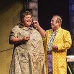 Edna in housecoat on the telephone, smiling as Wilbur smiles at her elbow, wearing yellow long coat with white polka dots, bright shirt and purple trousers