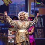 Motormouth Mabel in gold dress, singing with her arms raised in the air, the record store in the background