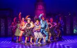 Tracy, Little Inez, Seaweed, Link and the Corney Collins cast dance on a geometric patterned floor with an ultra clutch hairspray bottle in purple shadow behind them