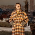 Gianni Schicchi (Dario Solari) in a tartan dressing gown, hands in pockets, bed in background